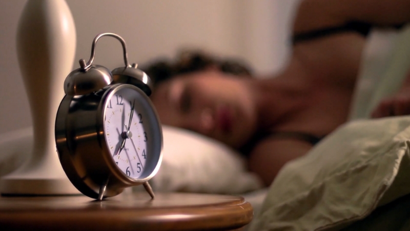 Budilnik na noćnom stočiću u prvom planu dok osoba mirno spava u krevetu u pozadini