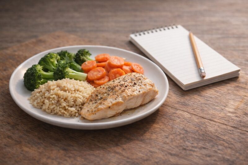 Zdrav obrok sa pečenim pilećim fileom, integralnim pirinčem, brokolijem i šargarepom na belom tanjiru na drvenom stolu